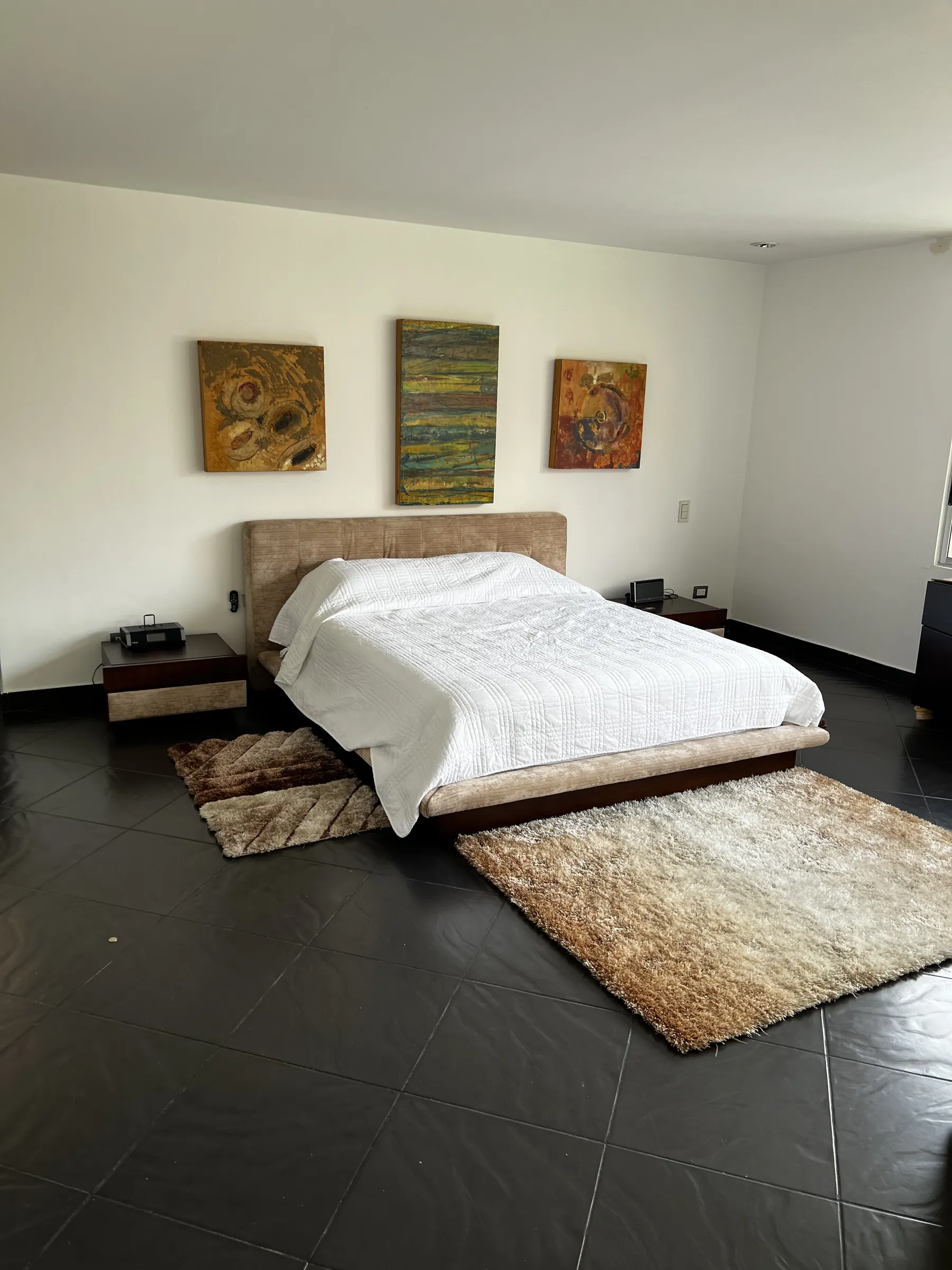 Bedroom with an upholstered platform bed, three framed canvases above the headboard and rugs on dark stone floors